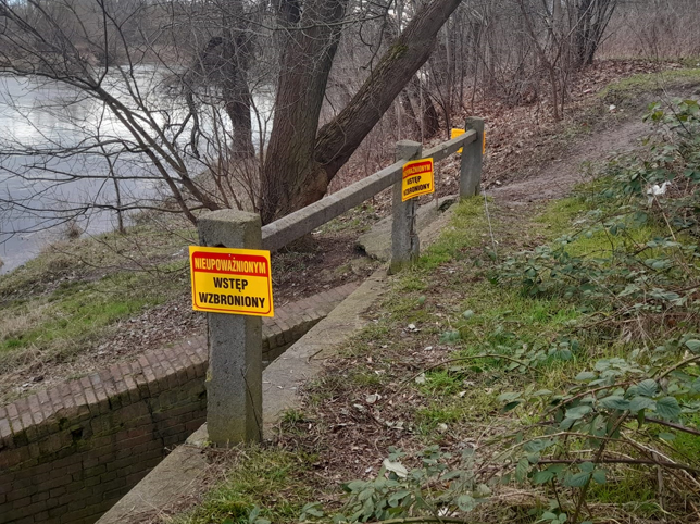 Zdjęcie pokazuje teren przy wejściu do kanału burzowego. Na zdjęciu widoczne są trzy tablice informujące o zakazie wstępu do kanału. Żółte tablice są przymocowane do betonowej barierki znajdującej się nad wejściem do kanału.